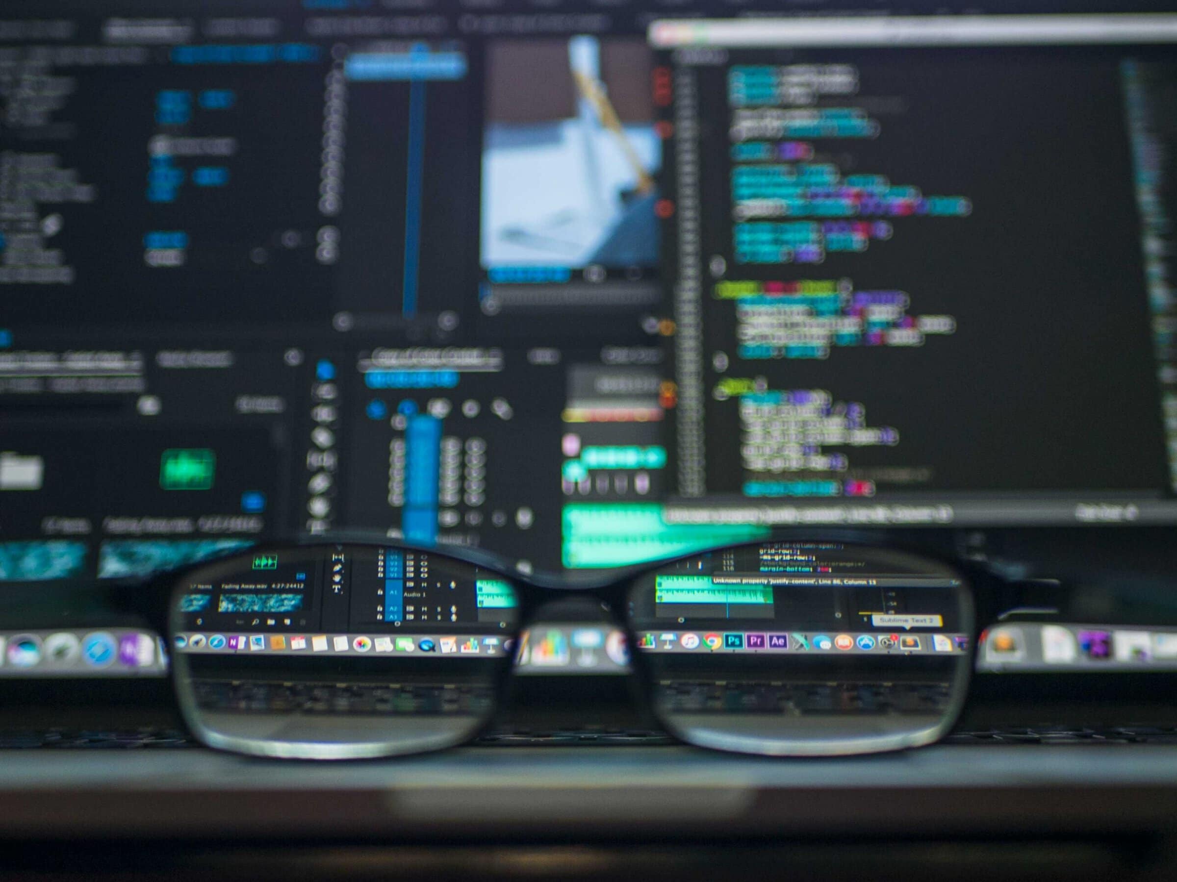 Black-rim eyeglasses in front of a laptop with a blurry screen. The laptop screen has windows open with various cybersecurity programs.
